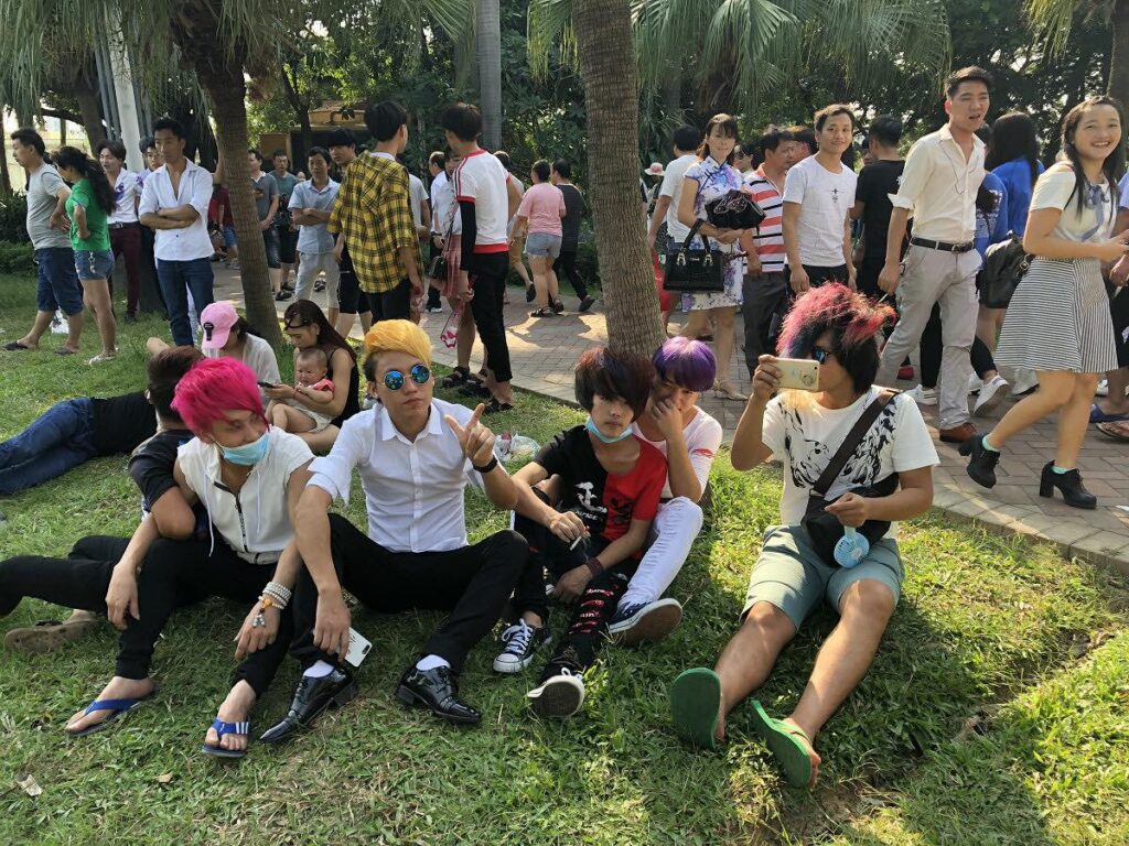 Four smarts with colorful hair sit on the grass in a park, while pedestrians in mainstream clothing walk behind them